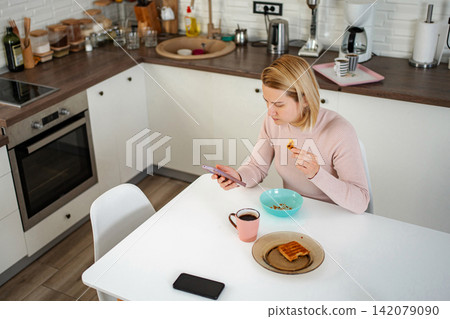 Woman using smartphone while eating breakfast and drinking coffee Woman using smartphone while eating breakfast and drinking coffee 142079090