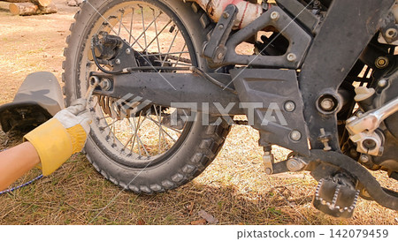 Close up of a person in a yellow-and-white work glove tightening a bolt on the rear wheel assembly of a dirty black motorcycle with a wrench Close up of a person in a yellow-and-white work glove tightening a bolt on the rear wheel assembly of a dirty black motorcycle with a wrench 142079459