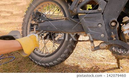 Close up of a person in a yellow-and-white work glove tightening a bolt on the rear wheel assembly of a dirty black motorcycle with a wrench Close up of a person in a yellow-and-white work glove tightening a bolt on the rear wheel assembly of a dirty black motorcycle with a wrench 142079460