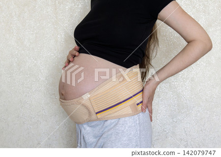 Close-up maternity photo of a pregnant woman wearing a supportive belly band and gently holding her baby bump. Concept of pregnancy care, comfort, health, and expecting motherhood. Close-up maternity photo of a pregnant woman wearing a supportive belly band and gently holding her baby bump. Concept of pregnancy care, comfort, health, and expecting motherhood. 142079747