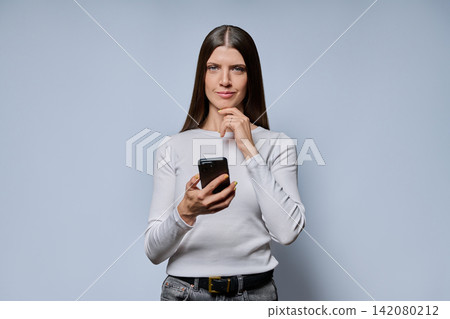 portrait of young woman holding smartphone and looking at camera on grey background portrait of young woman holding smartphone and looking at camera on grey background 142080212