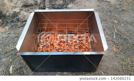 Close up of a two toned metal smoker box on dirt ground, filled with reddish wood chips beneath a metal grid perfect for outdoor smoking and campfire cooking. Close up of a two toned metal smoker box on dirt ground, filled with reddish wood chips beneath a metal grid perfect for outdoor smoking and campfire cooking. 142080231