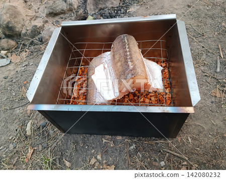 Top down view of a two toned metal smoker holding a large fish on a rack above reddish wood chips, placed on gravel and dirt with dried leaves in a camping setting. Top down view of a two toned metal smoker holding a large fish on a rack above reddish wood chips, placed on gravel and dirt with dried leaves in a camping setting. 142080232