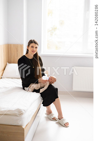 A young mother sits on a bed, gently feeding her newborn baby from a bottle in a bright, clean room 142080286