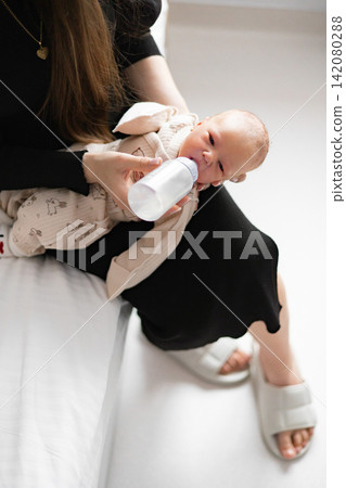 A caring mother gently feeds her newborn baby with a bottle while holding the infant close in her arms 142080288