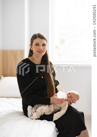 A young mother lovingly feeds her newborn baby from a bottle while sitting on a bed in a bright, minimalist room A young mother lovingly feeds her newborn baby from a bottle while sitting on a bed in a bright, minimalist room 142080291
