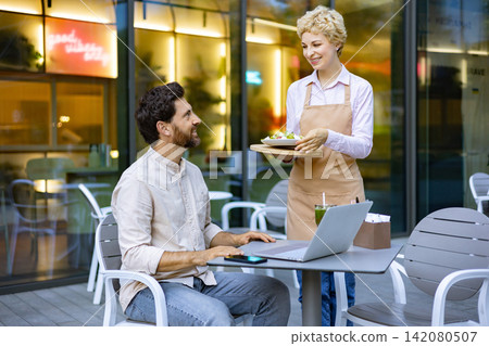 A smiling waiter in an apron serves a salad to a man sitting at a table with a laptop and phone outside a modern cafe A smiling waiter in an apron serves a salad to a man sitting at a table with a laptop and phone outside a modern cafe 142080507