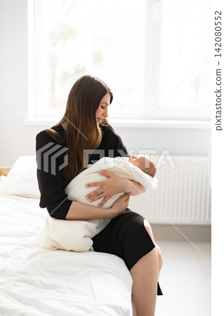 A young mother lovingly cradles her swaddled infant in her arms while sitting on a bed near a window A young mother lovingly cradles her swaddled infant in her arms while sitting on a bed near a window 142080552