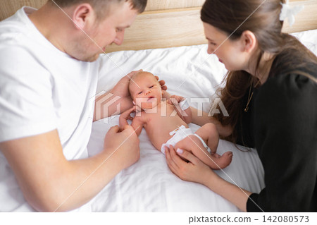 A loving family of three, a mother, father, and their newborn baby, share a tender moment together on a white bed 142080573
