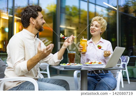 A man and woman are seated at a table outside a cafe, enjoying their meals and drinks while engaged in conversation A man and woman are seated at a table outside a cafe, enjoying their meals and drinks while engaged in conversation 142080719