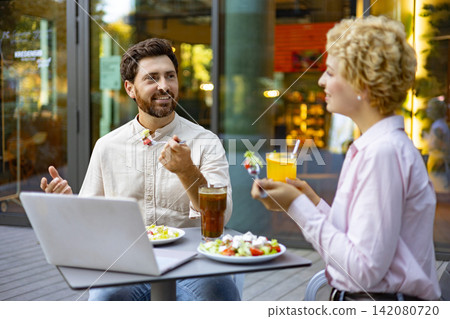 A man and a woman are having a pleasant conversation while eating salads and drinking beverages at an outdoor cafe table with a laptop A man and a woman are having a pleasant conversation while eating salads and drinking beverages at an outdoor cafe table with a laptop 142080720
