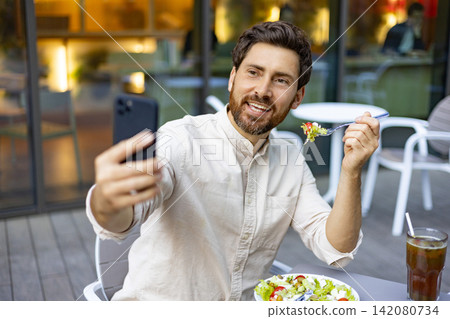 A smiling man with a beard takes a selfie with his smartphone while enjoying a fresh salad and a drink at an outdoor cafe A smiling man with a beard takes a selfie with his smartphone while enjoying a fresh salad and a drink at an outdoor cafe 142080734