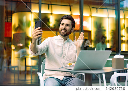 A smiling man with a beard is sitting at a cafe table with a laptop and holding up his smartphone to make a video call while waving hello A smiling man with a beard is sitting at a cafe table with a laptop and holding up his smartphone to make a video call while waving hello 142080738
