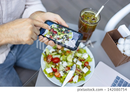 A person uses a smartphone to capture an image of a fresh, vibrant salad with feta cheese, tomatoes, and cucumbers, accompanied by a refreshing drink and a laptop A person uses a smartphone to capture an image of a fresh, vibrant salad with feta cheese, tomatoes, and cucumbers, accompanied by a refreshing drink and a laptop 142080754