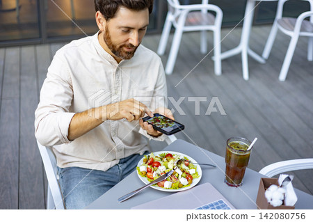 A man sits at an outdoor cafe table, using his smartphone to take a picture of his fresh Greek salad and iced beverage A man sits at an outdoor cafe table, using his smartphone to take a picture of his fresh Greek salad and iced beverage 142080756