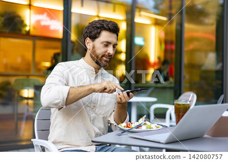 A man enjoys a meal outdoors while using his phone and working on a laptop at a cafe table A man enjoys a meal outdoors while using his phone and working on a laptop at a cafe table 142080757