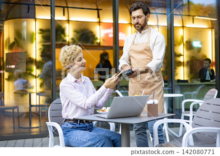 A woman uses her smartphone to make a contactless payment to a waiter at an outdoor cafe table with a laptop and salad A woman uses her smartphone to make a contactless payment to a waiter at an outdoor cafe table with a laptop and salad 142080771