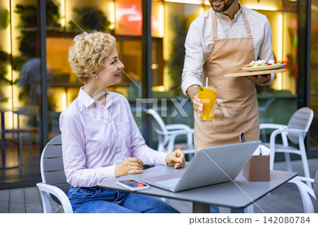 A smiling woman with blonde curly hair sits at a table with a laptop while a waiter in an apron serves her a refreshing orange drink and a salad A smiling woman with blonde curly hair sits at a table with a laptop while a waiter in an apron serves her a refreshing orange drink and a salad 142080784