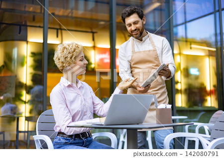 A smiling woman with blonde curly hair sits at a table with a laptop and discusses her order with a waiter holding a tablet A smiling woman with blonde curly hair sits at a table with a laptop and discusses her order with a waiter holding a tablet 142080791