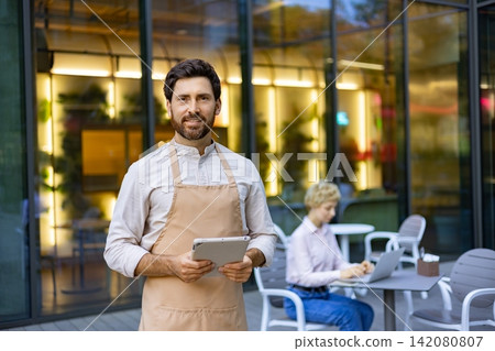 A smiling man wearing an apron holds a tablet while standing outside a restaurant with a blurred background of a patron working on a laptop A smiling man wearing an apron holds a tablet while standing outside a restaurant with a blurred background of a patron working on a laptop 142080807