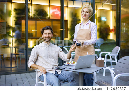 A man is paying with a credit card to a waitress at an outdoor cafe with a laptop on the table A man is paying with a credit card to a waitress at an outdoor cafe with a laptop on the table 142080817