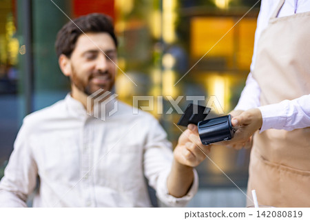 A man smiles while holding a credit card near a payment terminal held by a server wearing an apron A man smiles while holding a credit card near a payment terminal held by a server wearing an apron 142080819