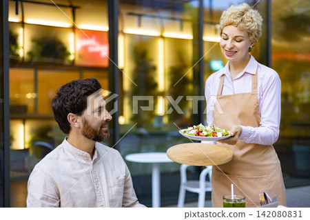 A smiling waitress in an apron presents a fresh salad to a man seated at an outdoor cafe table A smiling waitress in an apron presents a fresh salad to a man seated at an outdoor cafe table 142080831