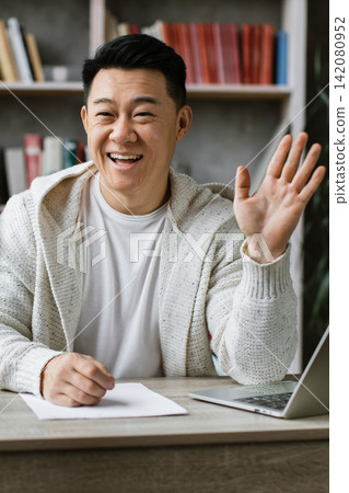A man smiles and waves at the camera while recording a video on his phone, with a laptop and books in the background A man smiles and waves at the camera while recording a video on his phone, with a laptop and books in the background 142080952