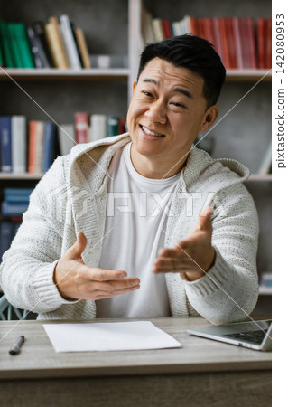 A man is sitting at his desk, talking to a camera set up on a tripod, with a laptop and bookshelves in the background 142080953