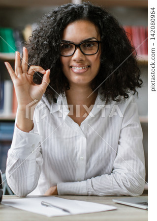 A young woman with curly hair and glasses smiles and gives an OK hand gesture while sitting at a desk with a laptop and headphones, ready for a video recording A young woman with curly hair and glasses smiles and gives an OK hand gesture while sitting at a desk with a laptop and headphones, ready for a video recording 142080964