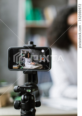 A woman with curly hair and glasses records herself on a smartphone for an online course while working on her laptop at a desk A woman with curly hair and glasses records herself on a smartphone for an online course while working on her laptop at a desk 142080972