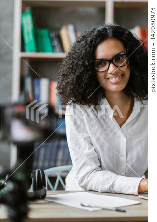A young woman with curly hair and glasses smiles while working on her laptop and recording a video with a camera in front of her 142080973