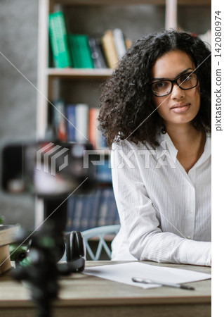 A woman with curly hair and glasses is recording a video with a camera while working on her laptop in a home office setting A woman with curly hair and glasses is recording a video with a camera while working on her laptop in a home office setting 142080974