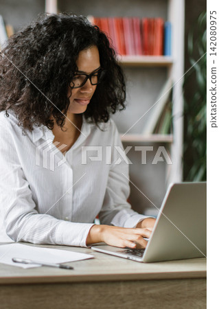 A woman with curly hair and glasses is typing on a laptop while a camera is set up in front of her, suggesting she is creating video content A woman with curly hair and glasses is typing on a laptop while a camera is set up in front of her, suggesting she is creating video content 142080975