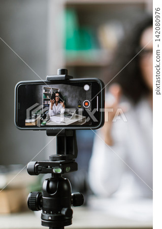 A woman with glasses and curly hair is recording herself on a smartphone mounted on a tripod, gesturing with her hands while sitting at a desk with a laptop and books A woman with glasses and curly hair is recording herself on a smartphone mounted on a tripod, gesturing with her hands while sitting at a desk with a laptop and books 142080976