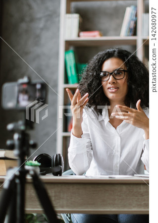A woman with glasses and curly hair is speaking animatedly while recording a video with her smartphone on a tripod in front of her laptop A woman with glasses and curly hair is speaking animatedly while recording a video with her smartphone on a tripod in front of her laptop 142080977