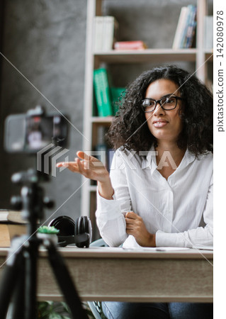 A woman with curly hair and glasses is recording a video with a camera on a tripod in her home office setup 142080978