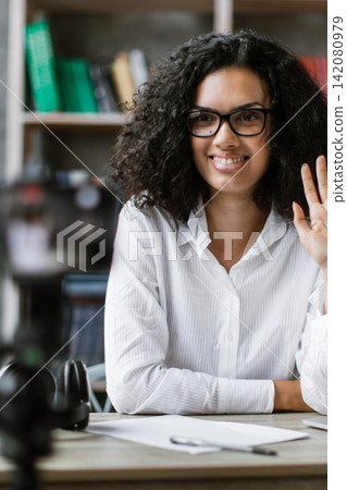 A young woman with curly hair and glasses smiles and waves hello while looking at the camera, positioned at her desk with a laptop and books 142080979