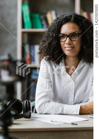 A young woman with glasses and curly hair sits at a desk in front of a bookshelf, preparing to record a video with a camera and laptop 142080980