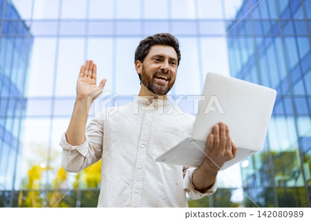 A smiling man with a beard holds a laptop and waves hello while standing outside a modern glass building, engaged in a virtual conversation 142080989