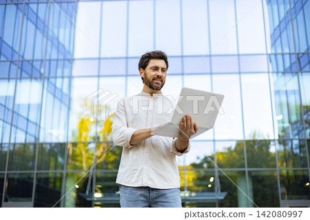 A smiling man with a beard stands outside a modern glass building holding a laptop and looking at the screen A smiling man with a beard stands outside a modern glass building holding a laptop and looking at the screen 142080997