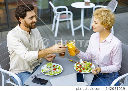 A man and woman enjoy a healthy meal and refreshing beverages while toasting glasses on an outdoor cafe patio A man and woman enjoy a healthy meal and refreshing beverages while toasting glasses on an outdoor cafe patio 142081019