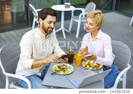 A man and woman are sitting at a table on a patio, enjoying salads and drinks while looking at their phones and a laptop A man and woman are sitting at a table on a patio, enjoying salads and drinks while looking at their phones and a laptop 142081020