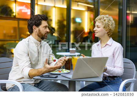 A man and a woman are engaged in a conversation while sitting at a table outside a cafe with a laptop and drinks A man and a woman are engaged in a conversation while sitting at a table outside a cafe with a laptop and drinks 142081030