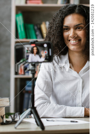A young woman with curly hair smiles and waves while recording a video on her smartphone placed on a tripod in her home office 142081219