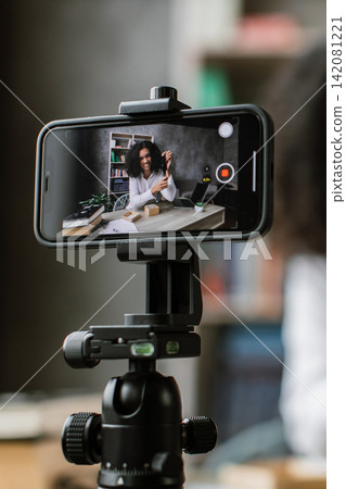 A young woman with curly hair is recording a video blog or tutorial using her smartphone mounted on a tripod, showcasing a product A young woman with curly hair is recording a video blog or tutorial using her smartphone mounted on a tripod, showcasing a product 142081221