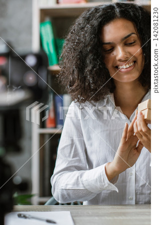 A young woman with curly hair smiles while holding a small cardboard gift box in front of a camera on a tripod, recording content A young woman with curly hair smiles while holding a small cardboard gift box in front of a camera on a tripod, recording content 142081230