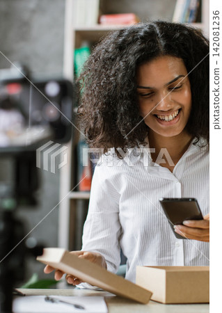 A smiling woman records herself opening a cardboard box while holding a smartphone, with bookshelves in the background A smiling woman records herself opening a cardboard box while holding a smartphone, with bookshelves in the background 142081236