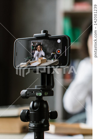 A young woman with headphones adjusts them while recording a video on her smartphone, which is mounted on a tripod 142081239
