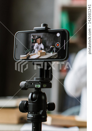A woman records herself on a smartphone mounted on a tripod, preparing to share content online A woman records herself on a smartphone mounted on a tripod, preparing to share content online 142081241